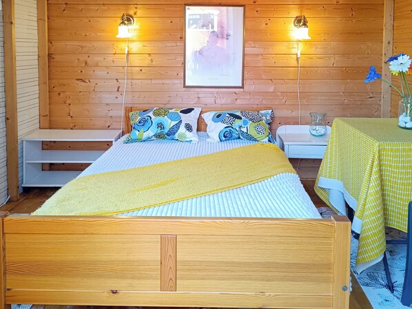 Una habitación de madera con una cama, dos mesitas de noche, una pequeña mesa con mantel amarillo y un cuadro en la pared.