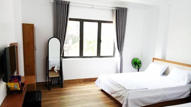A hotel room with two beds, a wooden floor, a window with curtains, a mirror, a green fan, and a TV on a wooden stand.