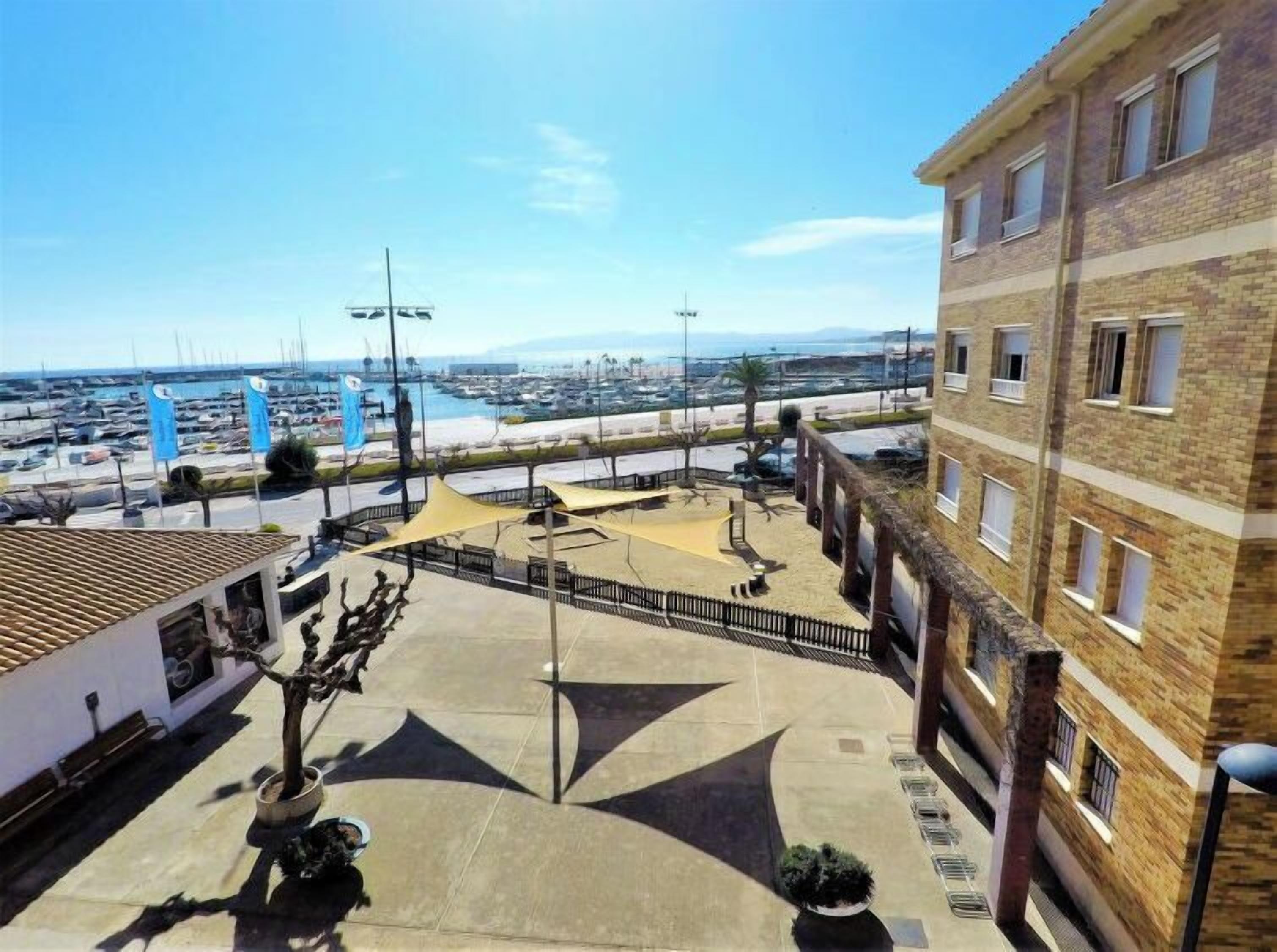 Une vue sur la côte, avec un port de plaisance, un bâtiment doté de nombreuses fenêtres et un espace extérieur ombragé avec un motif géométrique sur le sol.