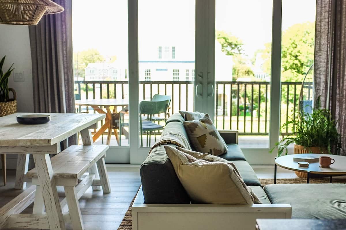 A cozy living room with a wooden table, a sofa with cushions, a small round table with a cup, and a view of the outside through large windows.