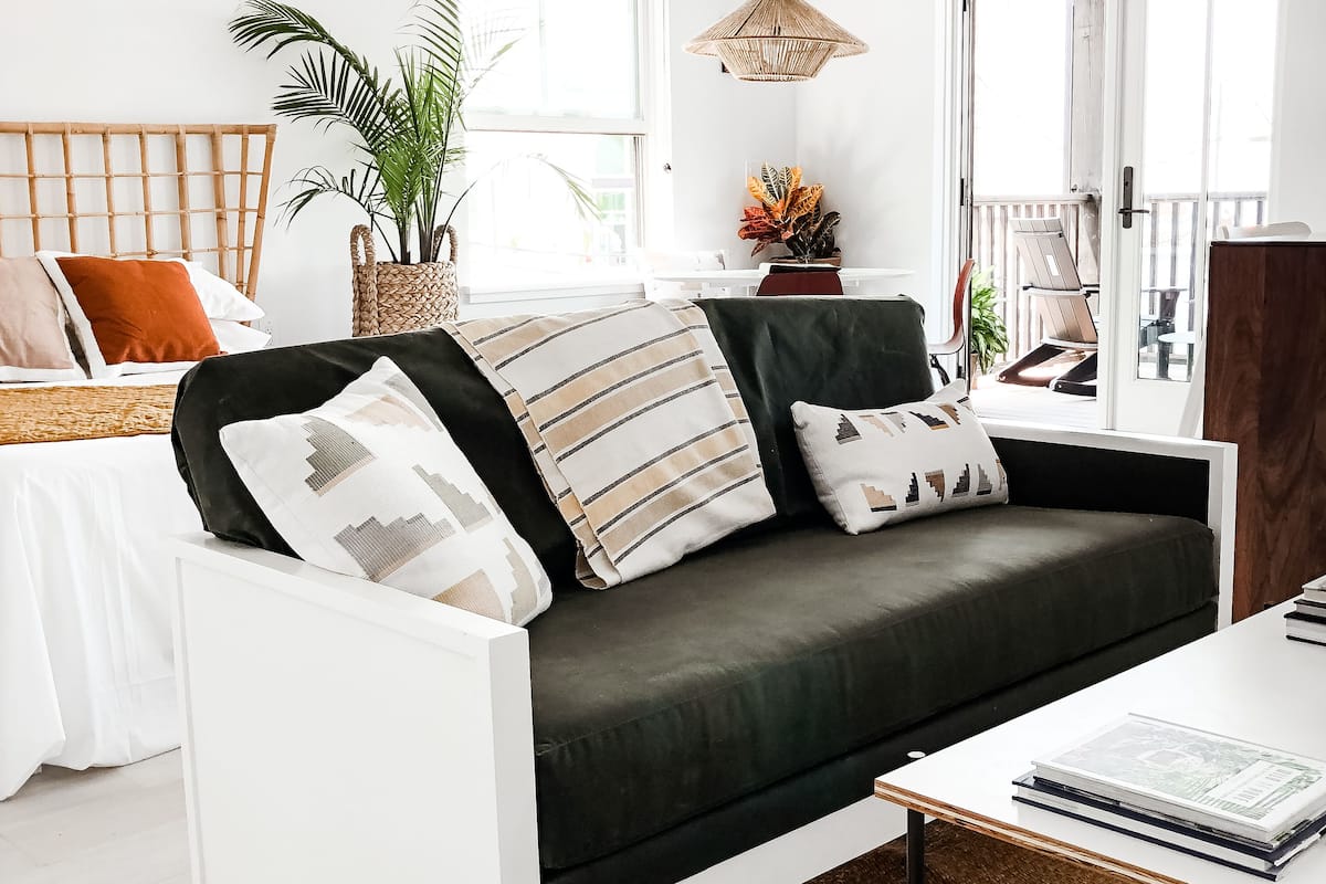 A modern living room with a dark green sofa, white armchairs, and a white coffee table. There is a wicker basket with plants, a wicker lamp, and a view of the balcony through the window.