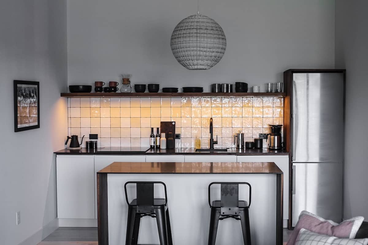 A modern kitchen with a central island, bar stools, a hanging light fixture, and a refrigerator.