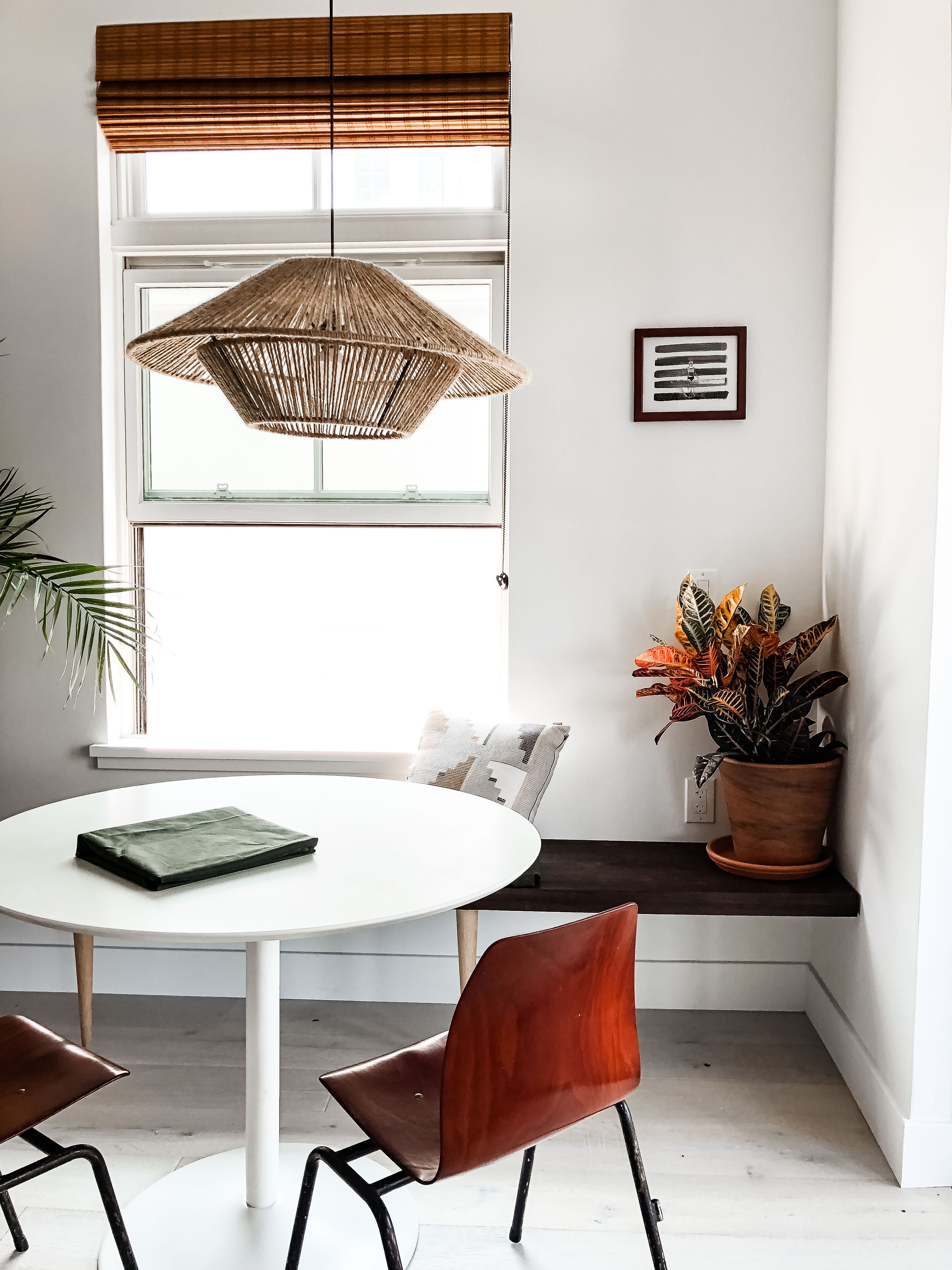 A dining area with a round table, two chairs, a hanging rattan light fixture, a window with blinds, a potted plant, and a framed picture on the wall.