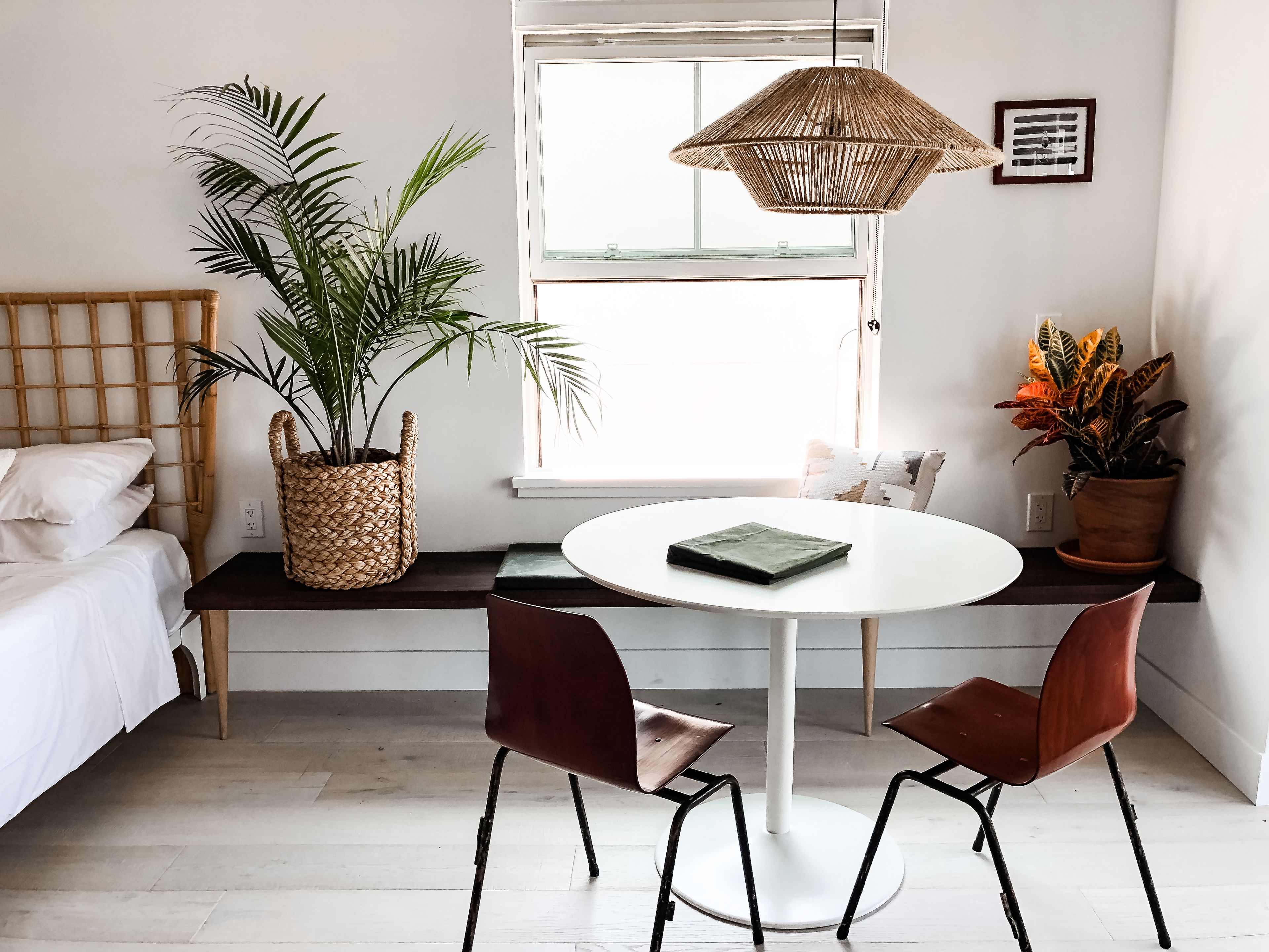 Una habitació amb un llit, una taula rodona amb dues cadires, un llum penjant i dues plantes en testos.