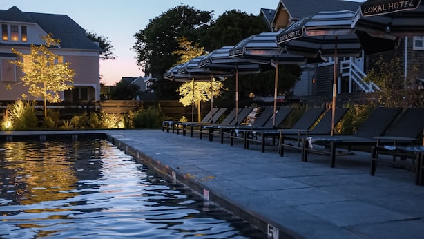 Un espace aménagé au bord de la piscine avec des chaises longues et des parasols, une enseigne indiquant « Hôtel local », et un reflet dans l’eau.