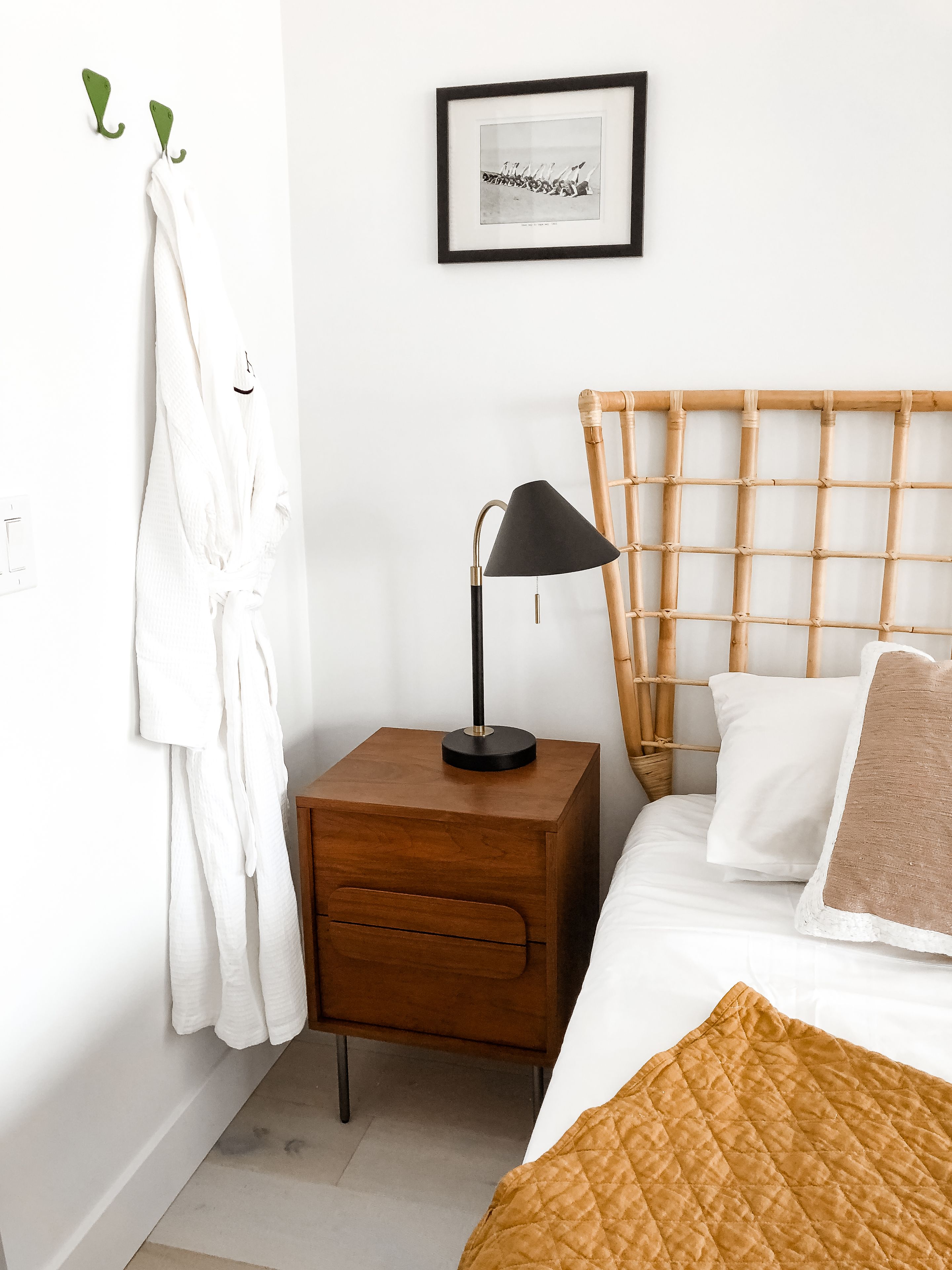 A neatly arranged bedroom with a wooden headboard, a bedside lamp, a framed picture on the wall, and a white robe hanging on hooks.