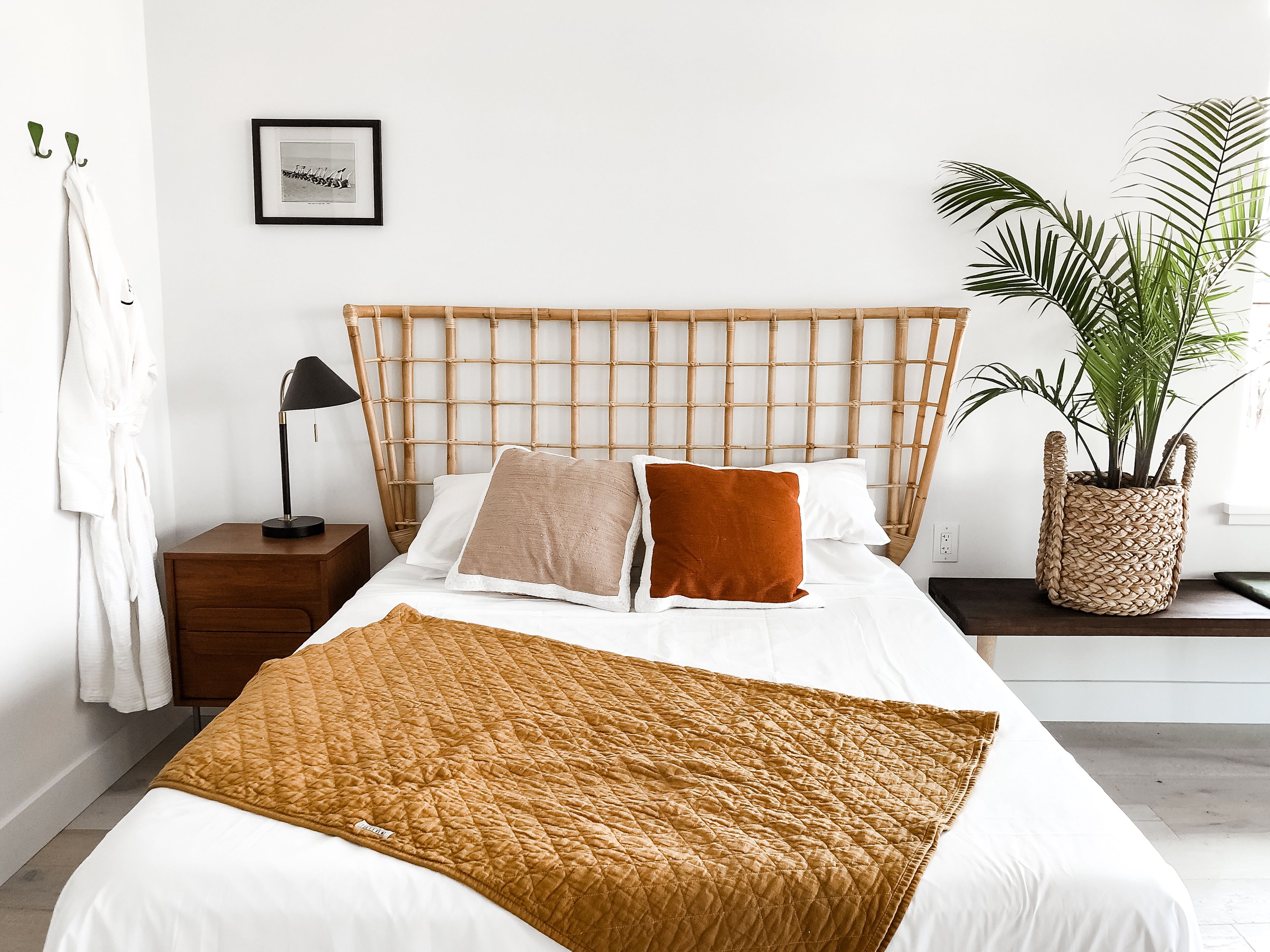 A neatly made bed with a brown quilt, a headboard with a woven design, a bedside lamp, a framed picture on the wall, and a potted plant.
