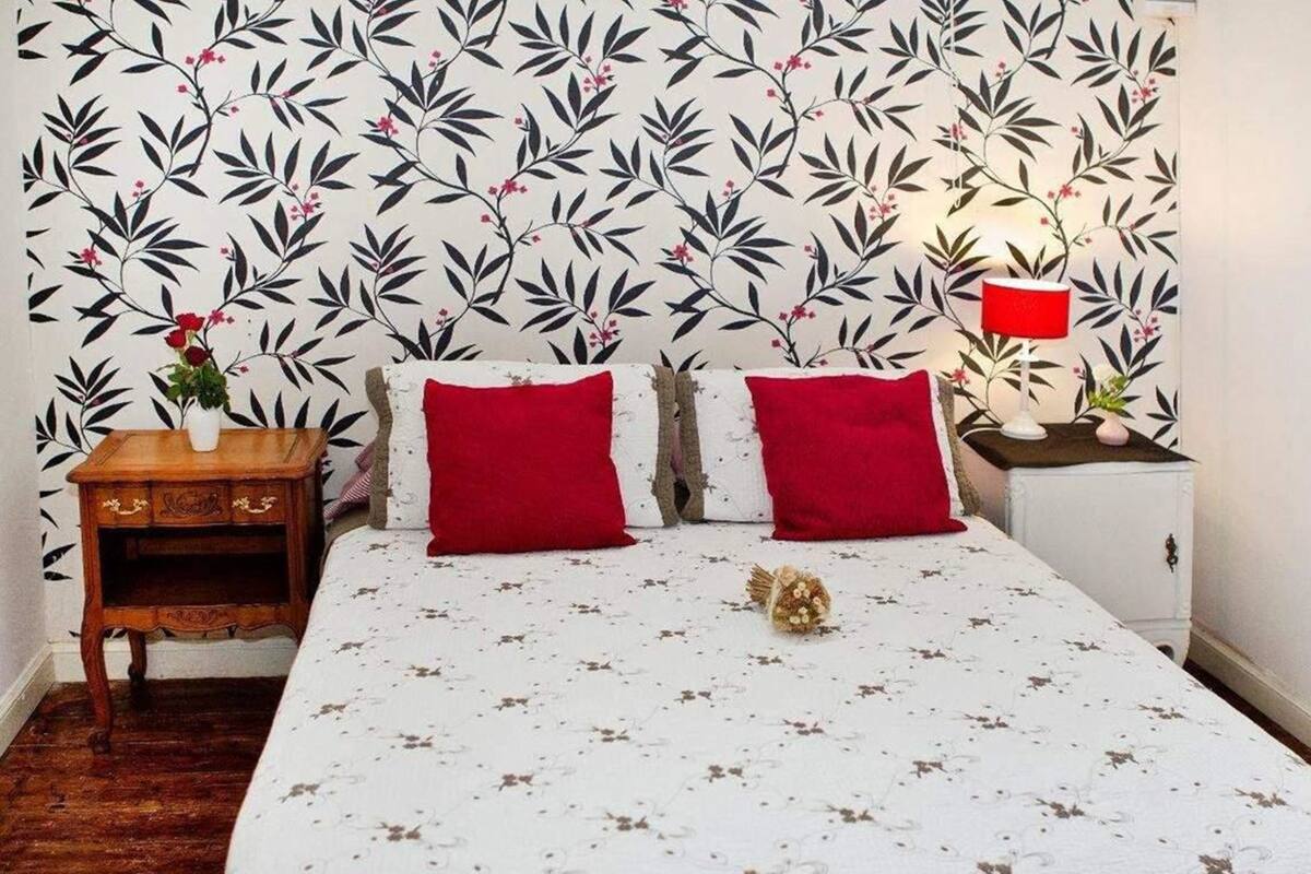 Un dormitorio con papel tapiz estampado con motivos florales, una mesita de noche de madera, una mesilla con una lámpara roja y una cama con dos almohadas rojas.