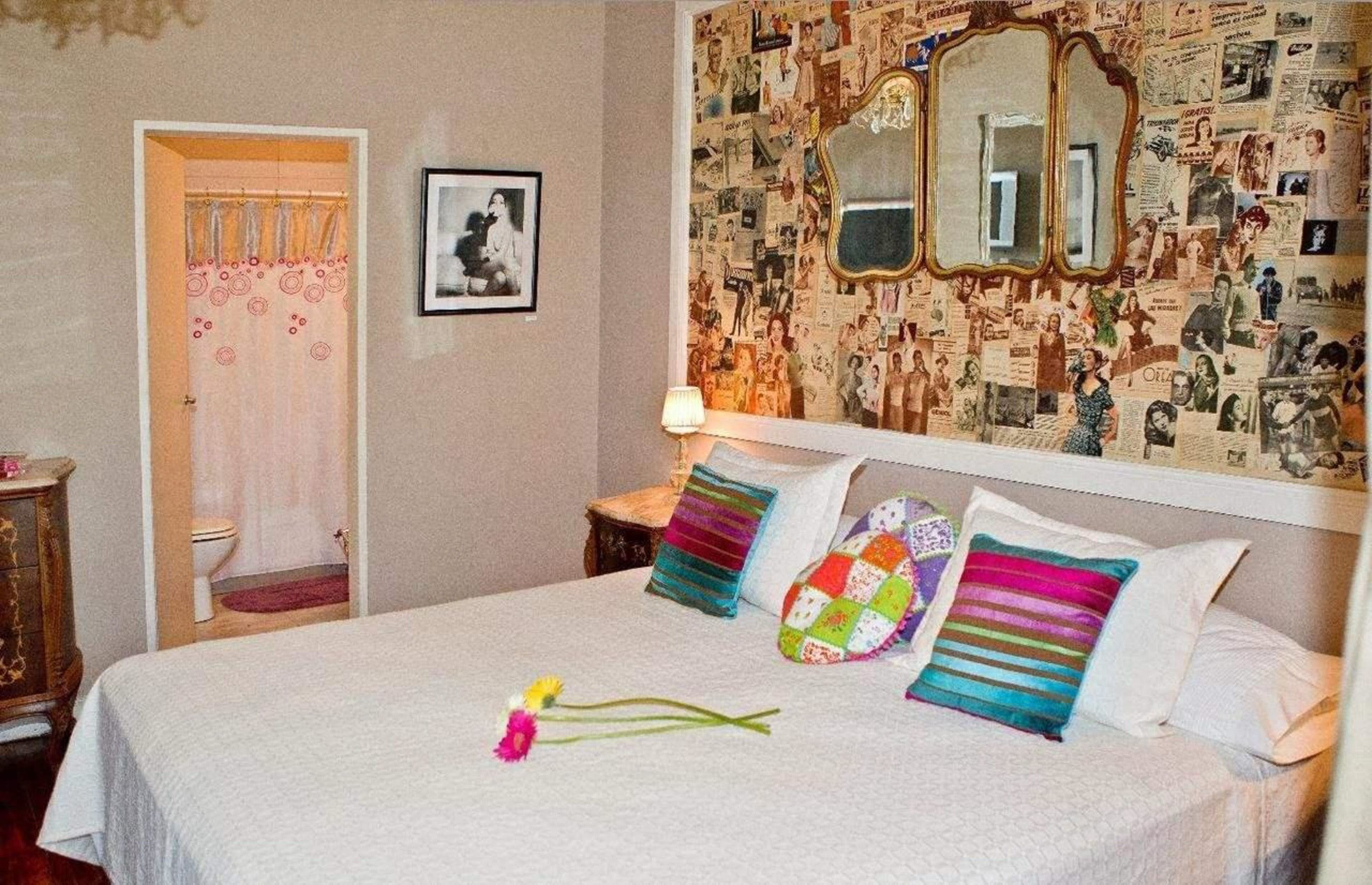 A bedroom with a bed, a bedside table, a mirror, and a wall covered with vintage photographs.