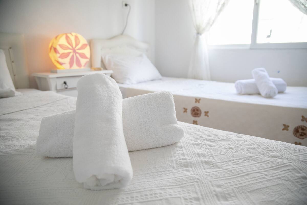 A neatly made bed with rolled white towels, a decorative wall lamp, and a window with curtains.