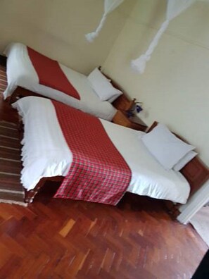 A room with two beds, each with white bedding and a red checkered blanket. There is a wooden floor and a nightstand with a lamp between the beds.