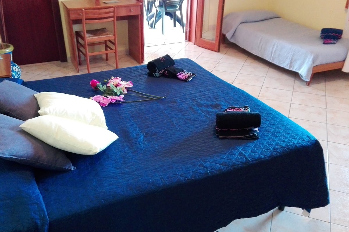 Quarto com cama, escrivaninha e cadeira. Há almofadas na cama e um vaso com flores.