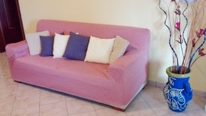 Un sofá rosa con varios cojines, un jarrón azul y blanco con ramas, y un suelo de baldosas.