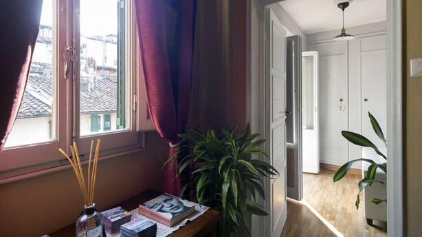 Ein Zimmer mit Fenster, einem Tisch mit Büchern und einer Pflanze, und Blick auf ein weiteres Zimmer mit Tür und Deckenleuchte.