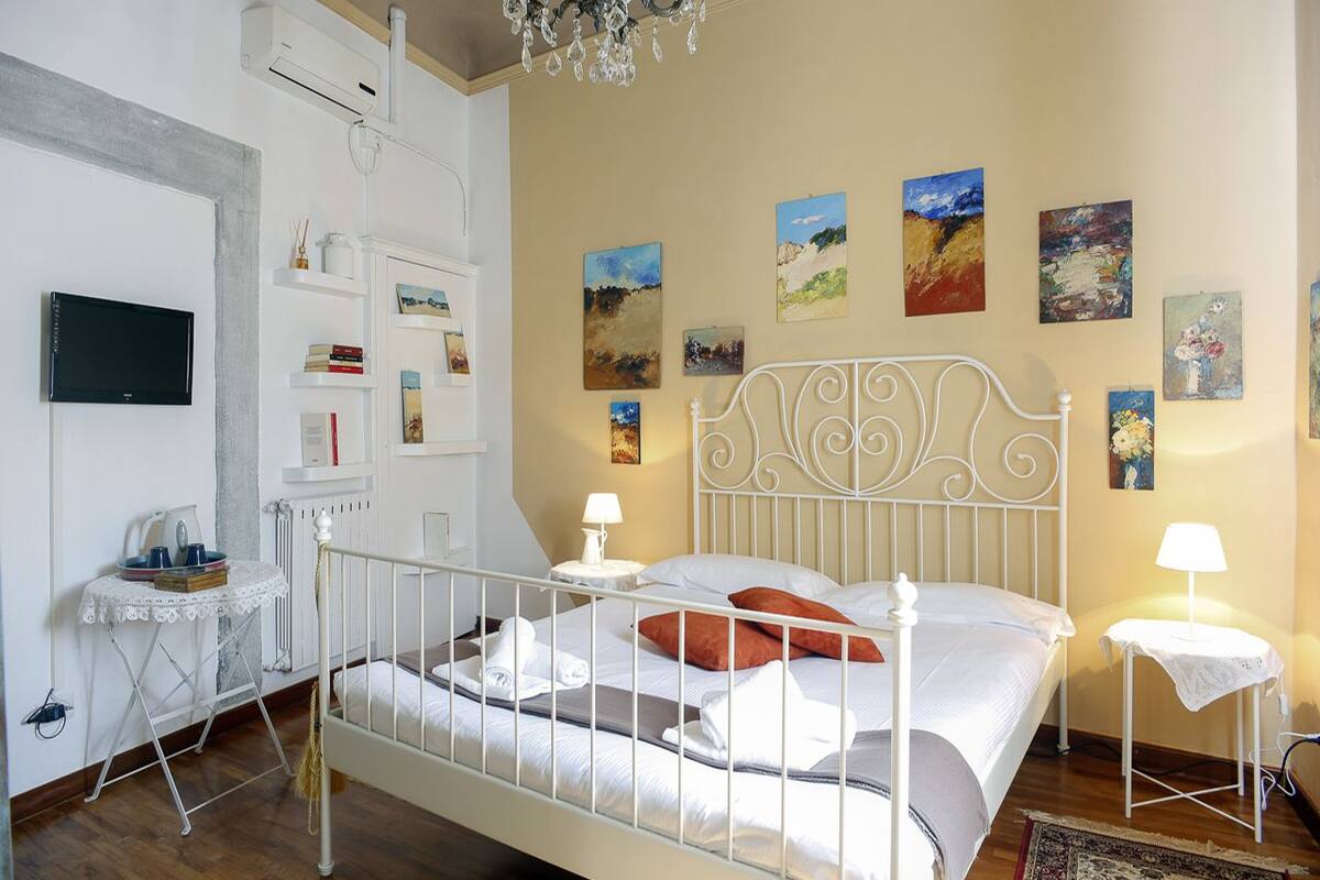 A bedroom with a white metal bed, a small bedside table with a lamp, a radiator, a wall-mounted TV, and various paintings on the wall.