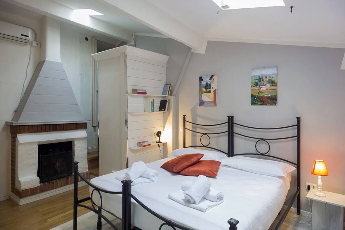 A bedroom with a bed, a fireplace, a nightstand with a lamp, and a painting on the wall.