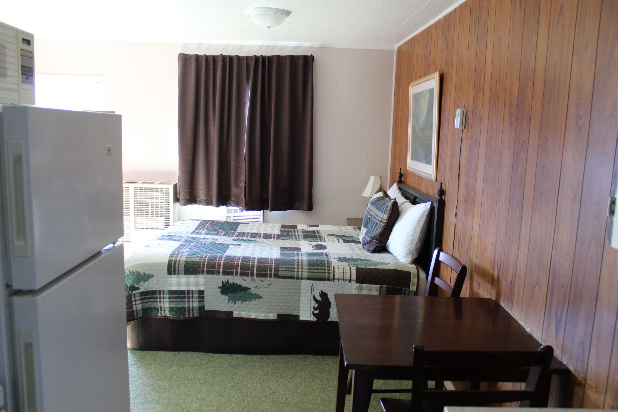 A room with a bed, a table, a chair, a refrigerator, a window with curtains, and a wall-mounted lamp.