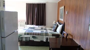 A room with a bed, a table, a chair, a refrigerator, a window with curtains, and a wall-mounted lamp.