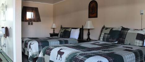 A hotel room with two beds, each with a quilted bedspread featuring a deer pattern. There is a window with curtains, a wall-mounted picture, and a nightstand with a lamp.