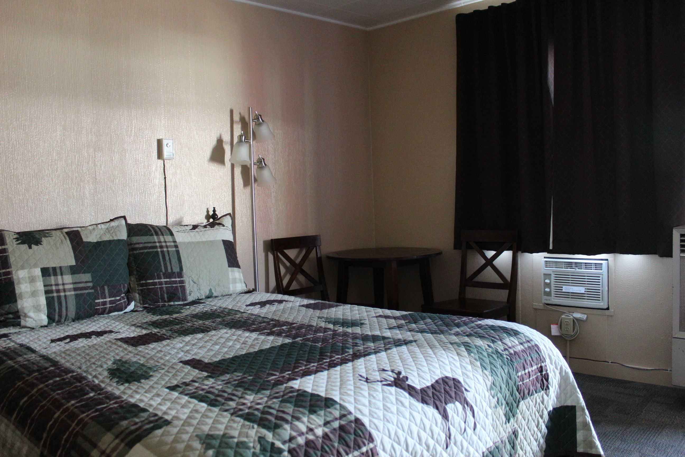 A hotel room with a large bed, a small table, a chair, a lamp, and an air conditioning unit.