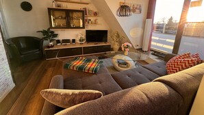 A modern living room with a sofa, a glass coffee table, a flat-screen TV, a bookshelf, and a large window with a view of the street. - 85 m² apartment, 3 rooms, kitchen, shower, 15 m² south-west facing balcony (Leipzig)