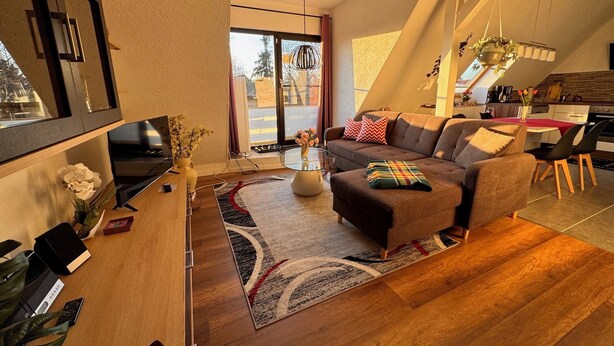 A modern living room with a sofa, coffee table, and a TV. There is a kitchen area with a dining table and chairs. - 85 m² apartment, 3 rooms, kitchen, shower, 15 m² south-west facing balcony (Leipzig)