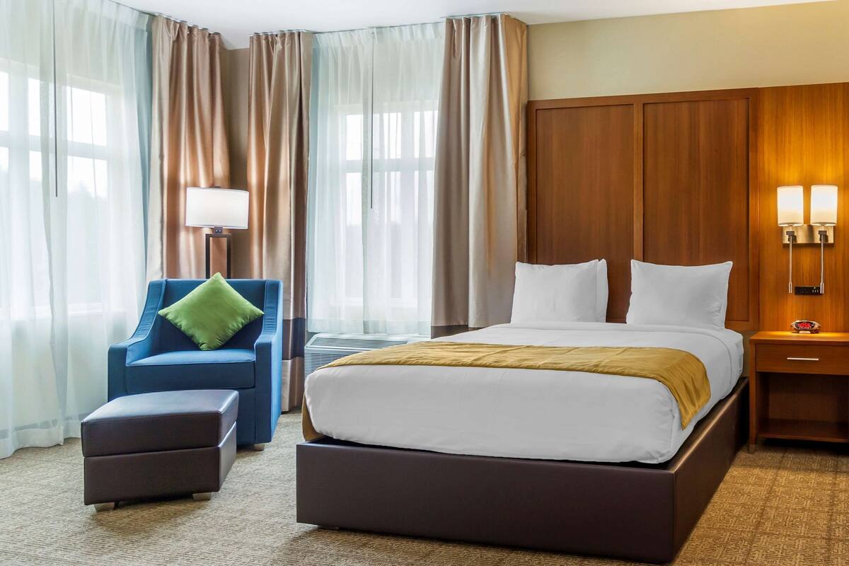 Une chambre d’hôtel avec un grand lit, un fauteuil bleu avec un coussin vert, un petit pouf, une table de chevet avec une lampe et une armoire en bois.