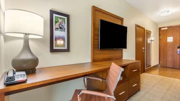 A hotel room with a wooden desk, a chair, a telephone, a television mounted on the wall, and a door.