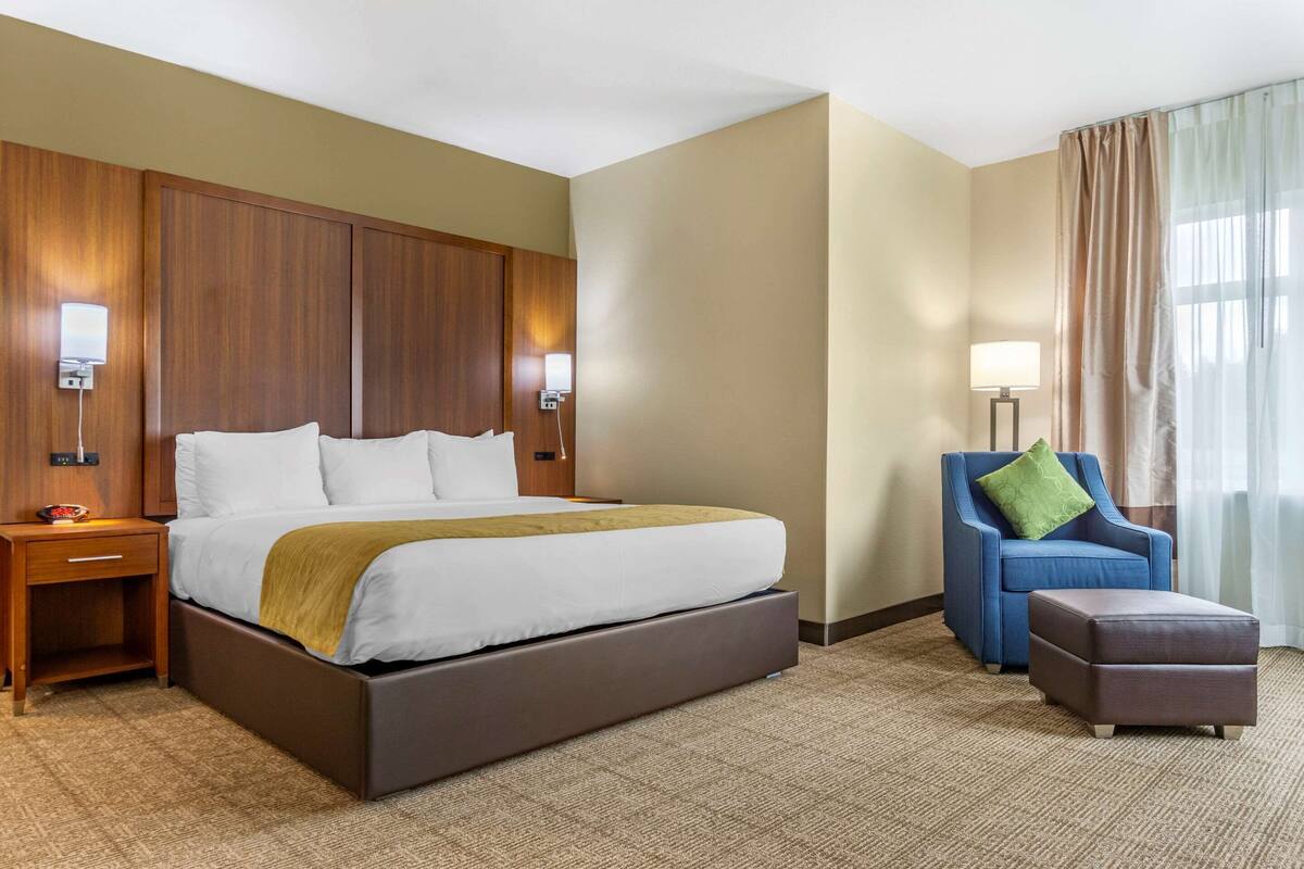 Une chambre d’hôtel avec un grand lit, une tête de lit en bois, un fauteuil bleu, une petite table et une fenêtre avec des rideaux.