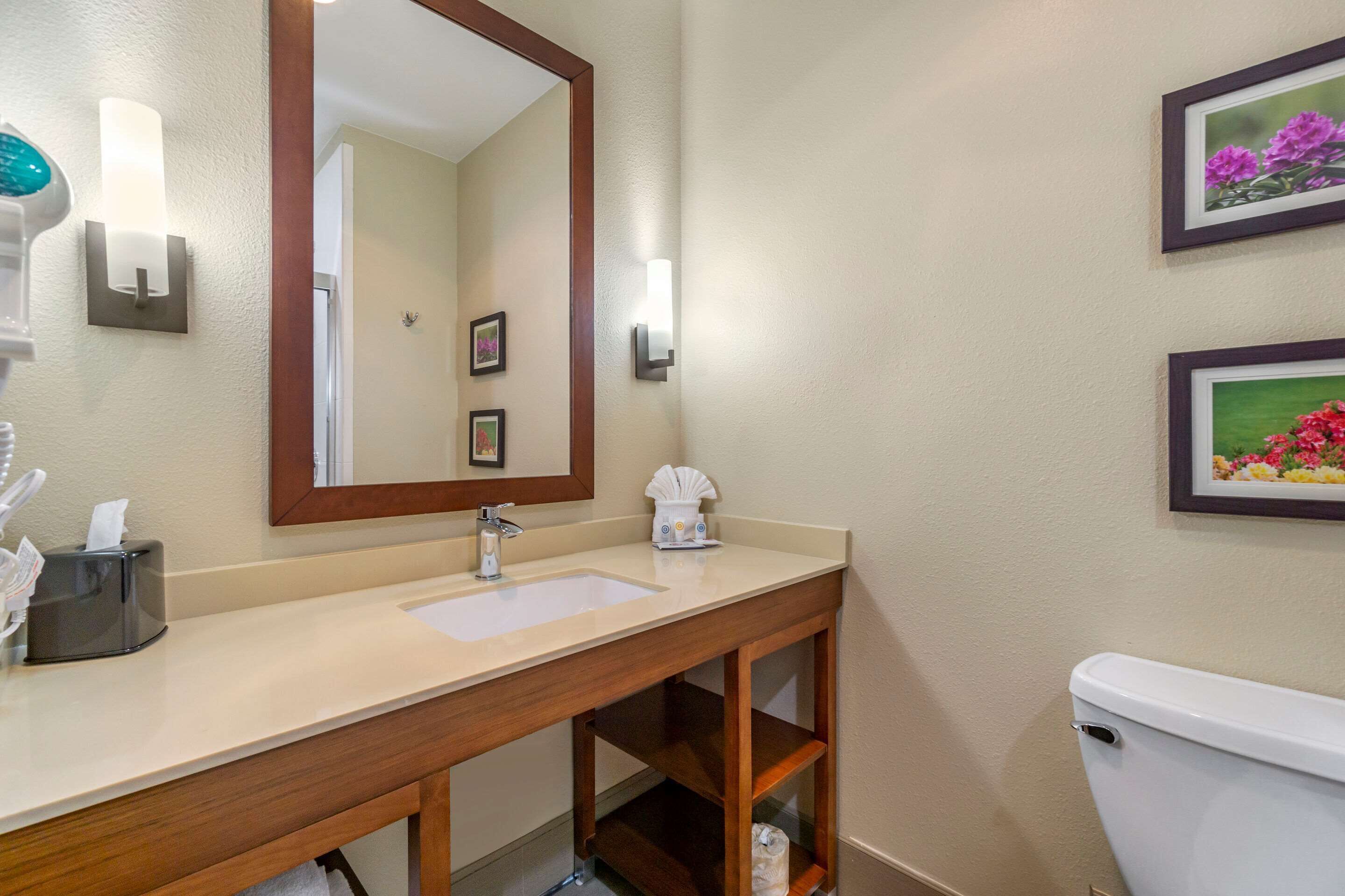 A bathroom with a wooden vanity, a sink, a mirror, and a toilet. There are framed pictures on the wall and a towel dispenser.