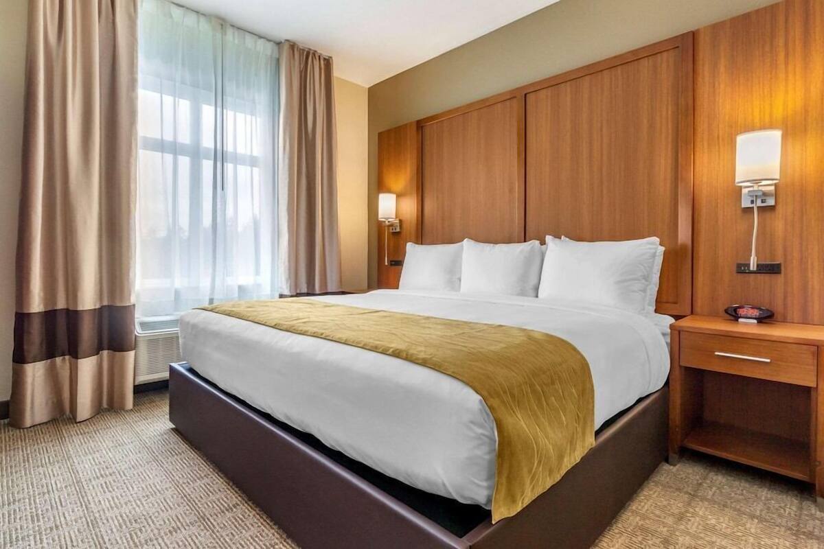Une chambre d’hôtel avec un grand lit, une tête de lit en bois, deux tables de chevet avec des lampes, une fenêtre avec des rideaux et un sol recouvert de moquette.