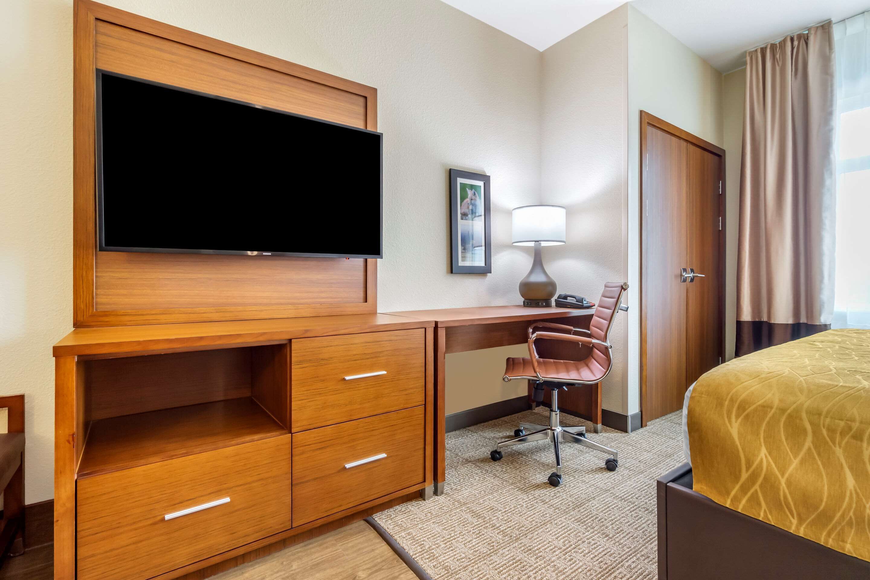 A hotel room with a wooden desk, a flat-screen TV, a chair, a bed, a lamp, and a door.