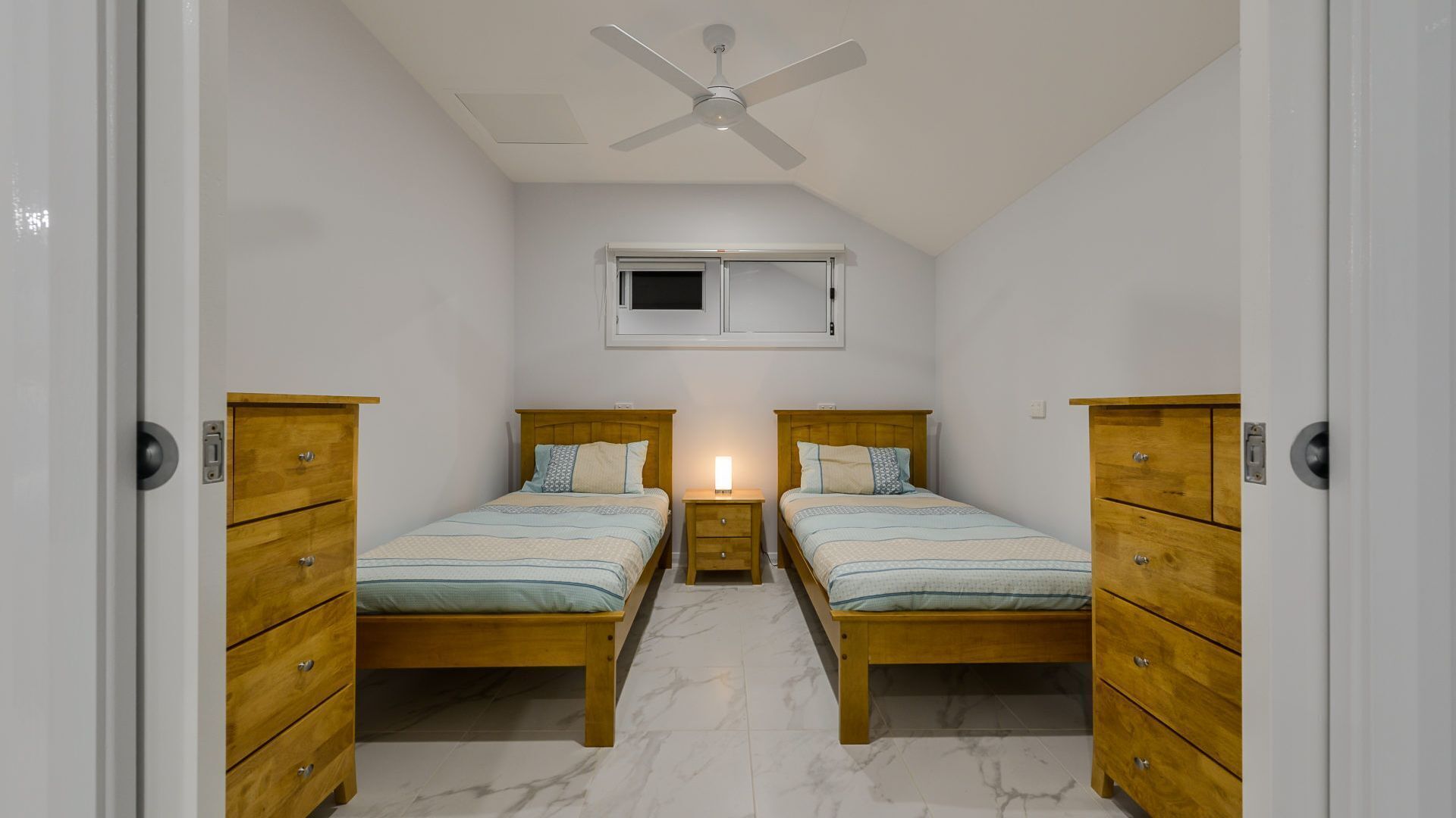 Quarto com duas camas, cómodas de madeira, ventoinha no teto, janela e mesa de cabeceira com abajur.