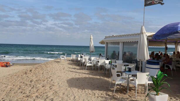 A beachside restaurant with outdoor seating, a flag, and the ocean in the background.