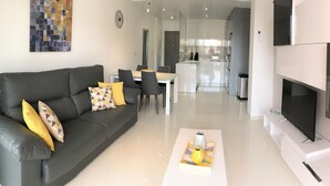 A modern living room with a grey sofa, a flat-screen TV, and a dining area with a white table and chairs.