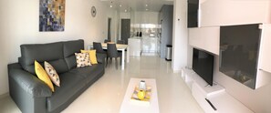 Un salon moderne avec un canapé gris, un téléviseur à écran plat et un coin repas comprenant une table et des chaises blanches.