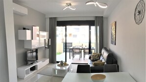 A modern living room with a flat-screen TV, a sofa, a dining table with chairs, and a large window with curtains.