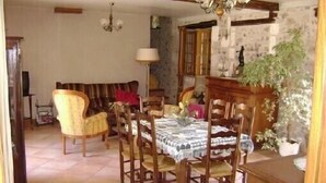 Uma sala de jantar com mesa de madeira, cadeiras e um candelabro. Há um sofá e um abajur ao fundo.
