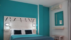 A bedroom with teal walls, a wooden headboard with bird silhouettes, a bed with teal bedding, bedside tables with lamps, a small bedside table, and an air conditioning unit. - Hotel El Pino (Teror)