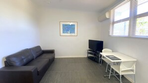 A small, modern room with a dark leather sofa, a flat-screen TV, a white desk, and a framed artwork on the wall. - Getaway Villas Unit 38-11 - comfort on a budget (EXMOUTH)