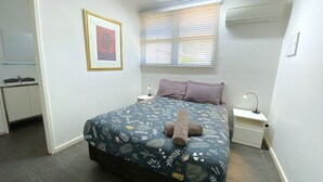 A bedroom with a bed, a bedside table, a nightstand, a window with blinds, a framed picture on the wall, and a small cabinet.