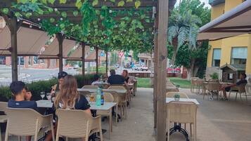Un café al aire libre con mesas y sillas bajo una pérgola de madera con plantas colgantes.