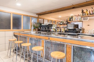 Una zona de bar con mostrador de mármol, taburetes de madera, una cafetera y estantes con botellas.
