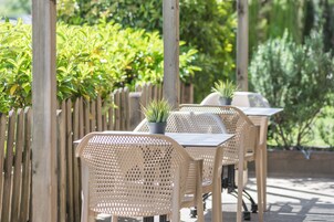 Un patio al aire libre con sillas y mesas de mimbre, rodeado por una valla de madera y abundante vegetación.