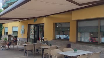 Uma área externa com mesas e cadeiras de vime, um alpendre coberto e um edifício amarelo com sacadas.