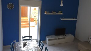 Et værelse med blå vægge, en hvid dør, et spisebord med stole, et fjernsyn på et stativ og en lille reol med bøger.
