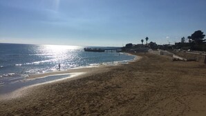 En strand med en mole, der strækker sig ud i vandet, en klar himmel og nogle få personer, der går langs kysten.