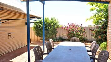A covered patio area with a table and chairs, surrounded by a wooden fence and flowering bushes.