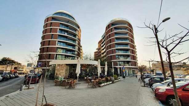 A modern building with a curved facade, outdoor seating area, and a parking lot. - Design Apt Parking Included 2BDR TL30 (Tel Aviv)