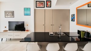 A modern kitchen with a black countertop, white chairs, a built-in oven, and a large mirror with an orange frame. - Design Apt Parking Included 2BDR TL30 (Tel Aviv)